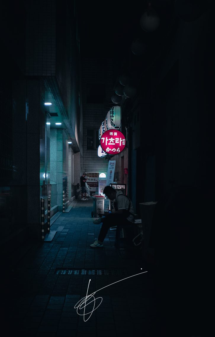 a shot of a dark alleyway, with a person smoking in the foreground and one staring at the phone in the background